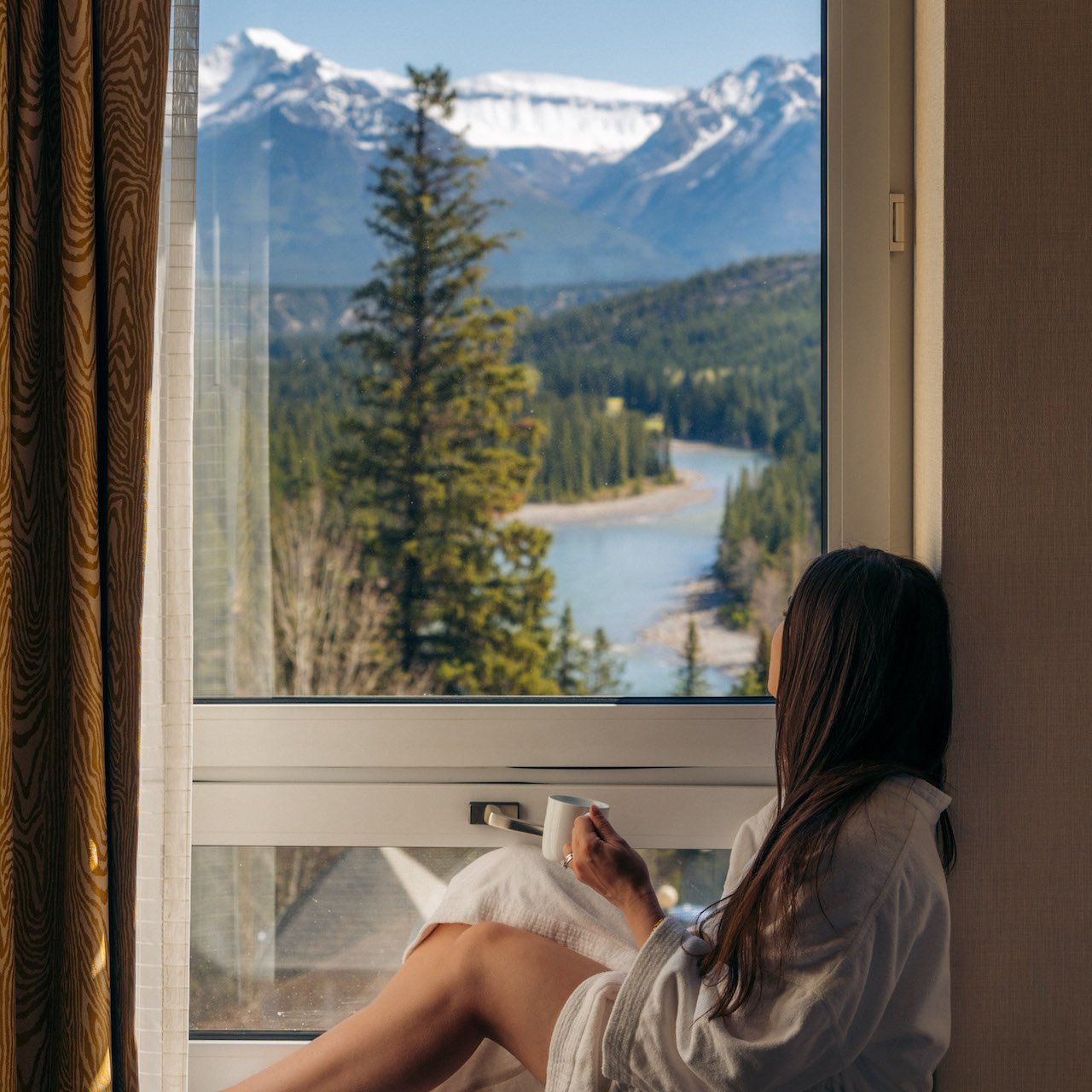 girl in bathrobe sitting on hotel room windowsill with coffee in hand facing mountain view
