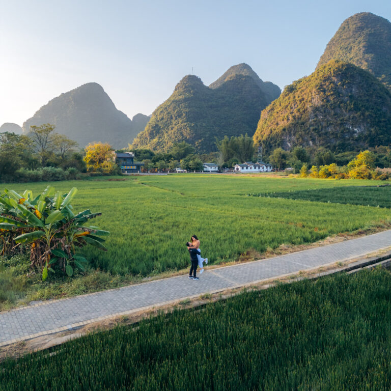 Jason and Lauren embracing along diagonal path amidst Yangshuo, Guilin valley, with lush rice fields and limestone mountains, golden with soft morning glow