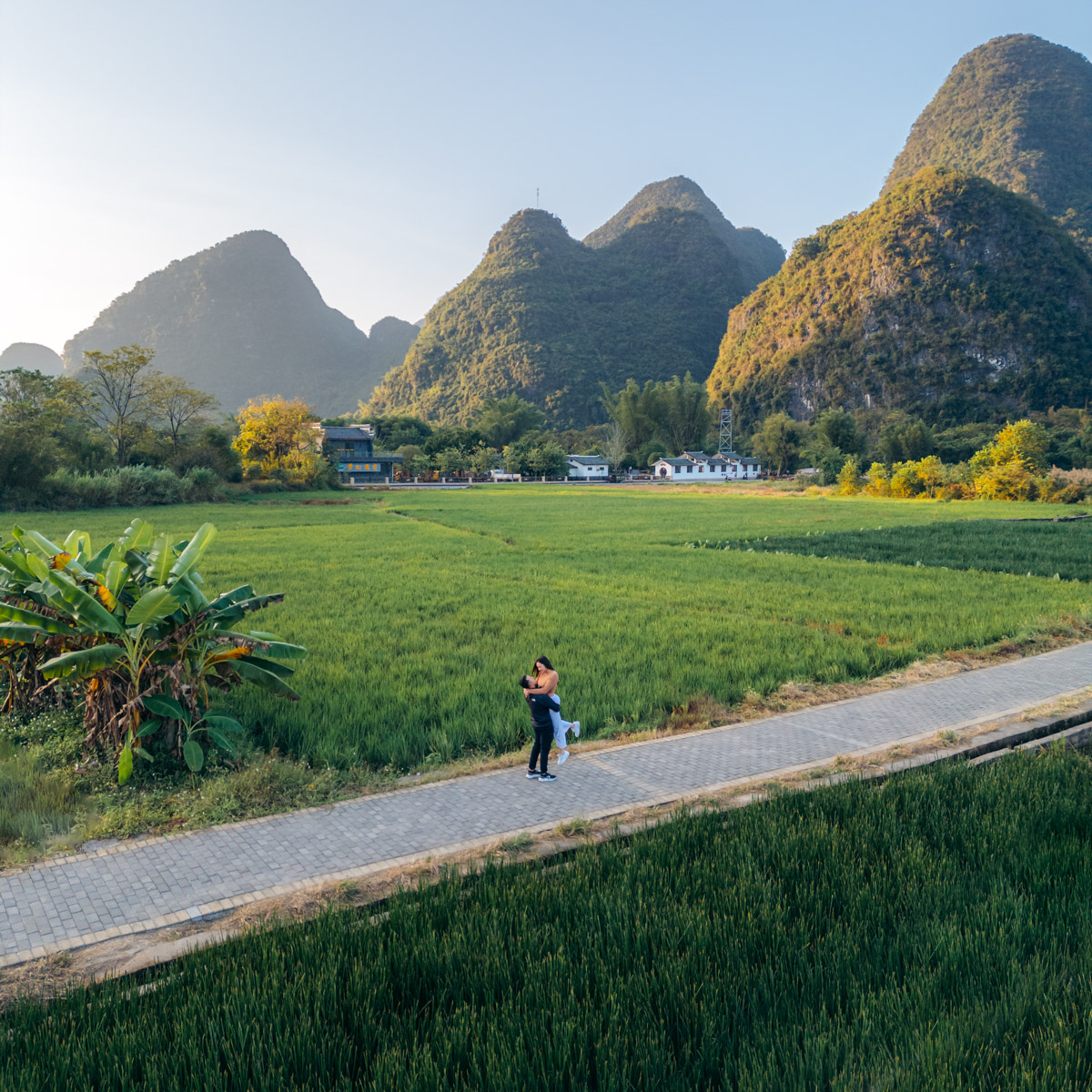 Jason and Lauren embracing along diagonal path amidst Yangshuo, Guilin valley, with lush rice fields and limestone mountains, golden with soft morning glow